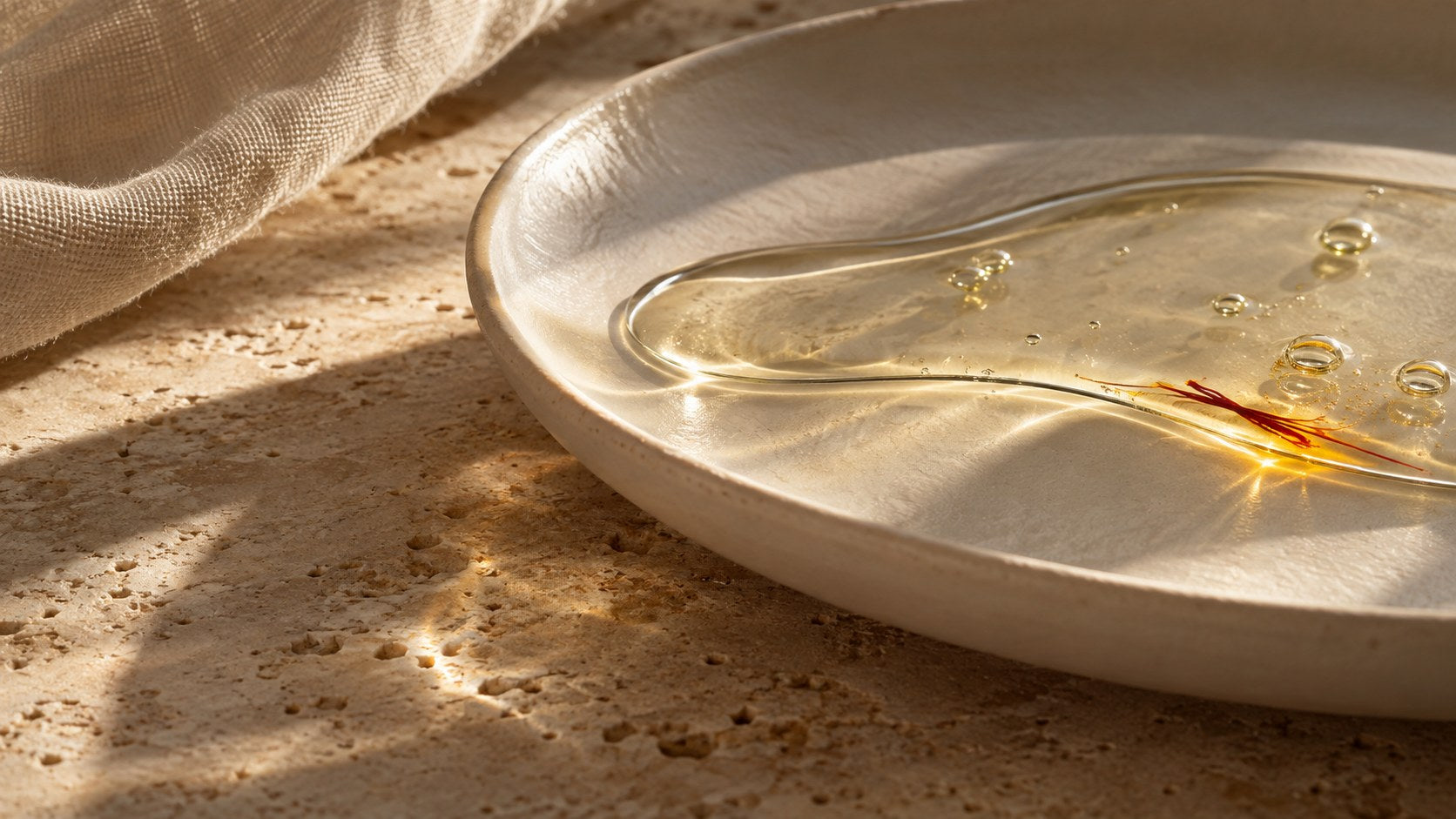 Clear oil, saffron threads and tiny air bubbles catching warm light in a matte porcelain dish.