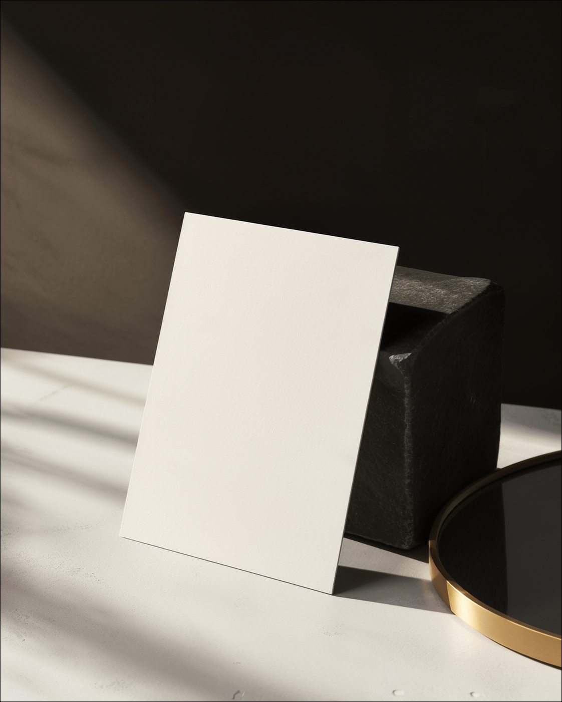 A blank ivory card leaning against a warm-charcoal stone block beside a gold-edged mirror.