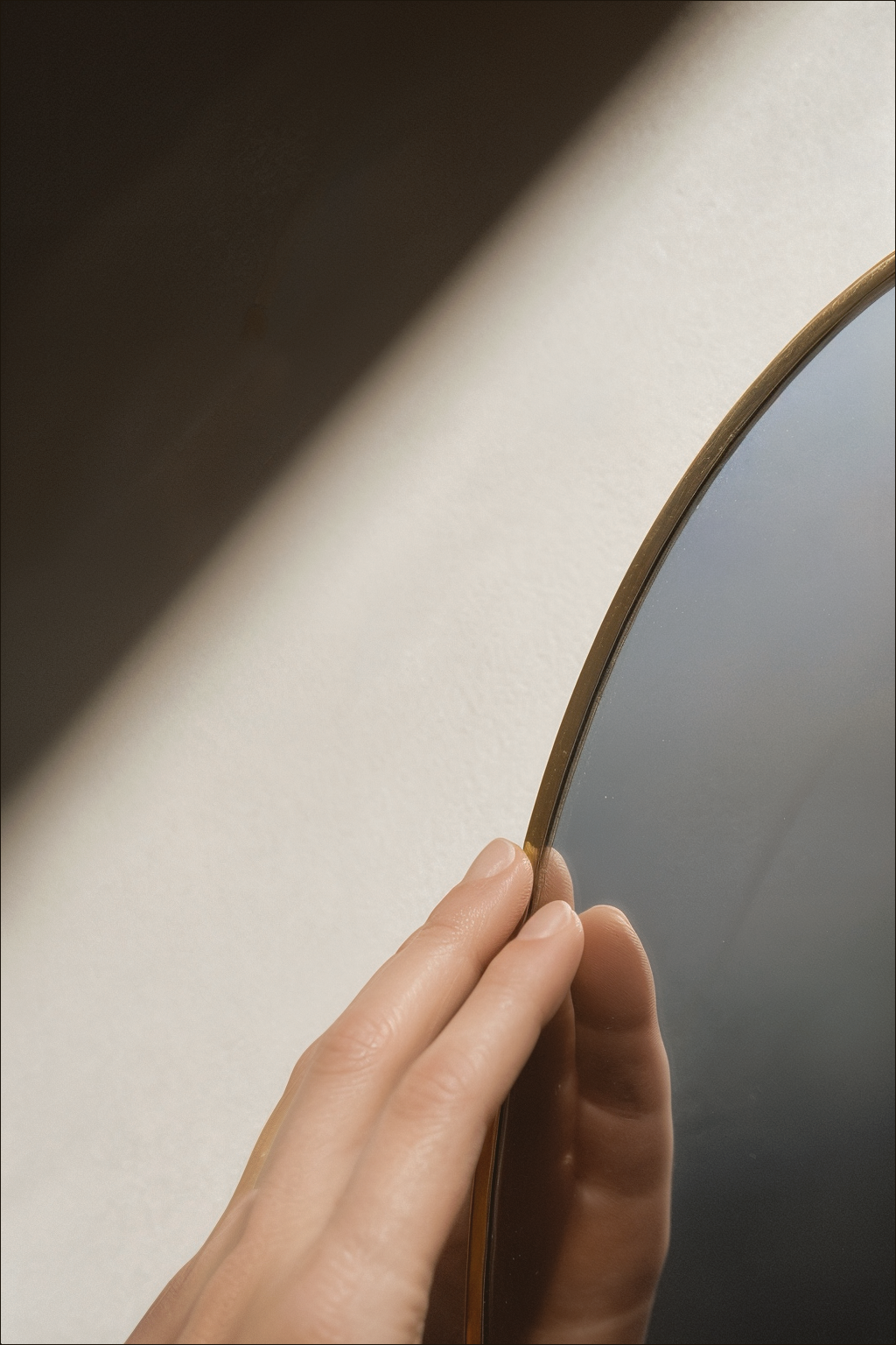 Natural fingertips touching the edge of a gold-rimmed mirror in ivory light.