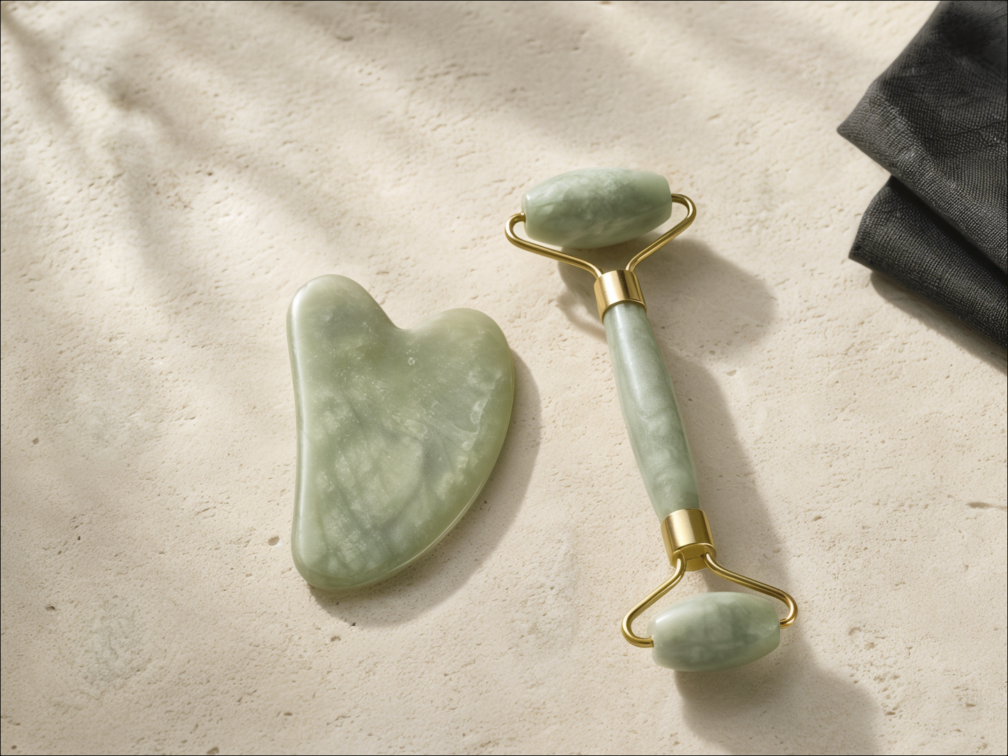 A pale jade gua sha and matching facial roller resting side by side on warm ivory stone.